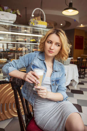 Blonde (with dark roots) caucasian woman in casual summer outfit at the cafe. Gray dress and jeans jacket. Woman got natural day makeup and curly hairstyle. Outdoorsの写真素材