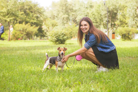 Cute beautiful slim caucasian brunette woman in casual outfit (skirt, shirt and jeans jacket) on a walk in park in summer day with her adorable jack russell dog. They full of joy, smiling and have funの写真素材
