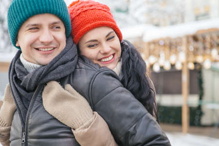 Cheerful smiling caucasian couple of man and woman having fun on european street walk. Casual outfit: jeans, jackets and hats. Winter cold weather with snow. Decorated Christmas tree on a backgroundの写真素材