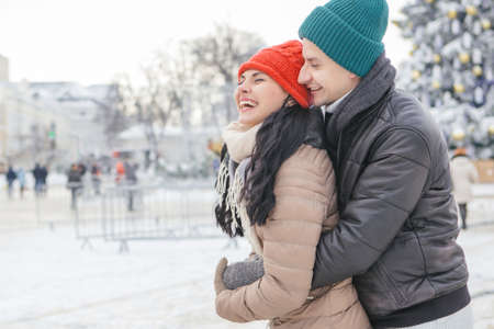 Cheerful smiling caucasian couple of man and woman having fun on european street walk. Casual outfit: jeans, jackets and hats. Winter cold weather with snow. Decorated Christmas tree on a backgroundの写真素材