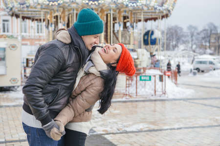 Cheerful smiling caucasian couple of man and woman having fun on european street walk. Casual outfit: jeans, jackets and hats. Winter cold weather with snow. Decorated Christmas tree on a backgroundの写真素材