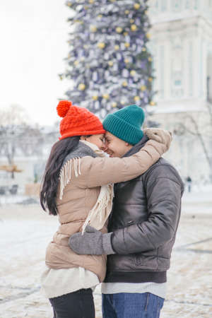 Cheerful smiling caucasian couple of man and woman having fun on european street walk. Casual outfit: jeans, jackets and hats. Winter cold weather with snow. Decorated Christmas tree on a backgroundの写真素材