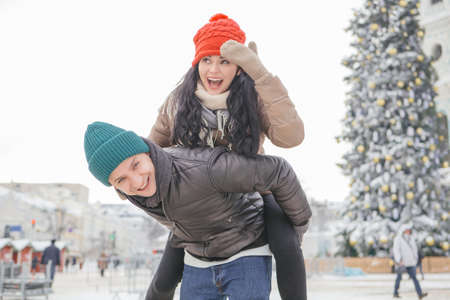 Cheerful smiling caucasian couple of man and woman having fun on european street walk. Casual outfit: jeans, jackets and hats. Winter cold weather with snow. Decorated Christmas tree on a backgroundの写真素材