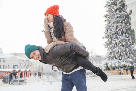 Cheerful smiling caucasian couple of man and woman having fun on european street walk. Casual outfit: jeans, jackets and hats. Winter cold weather with snow. Decorated Christmas tree on a backgroundの写真素材