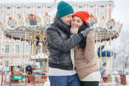 Cute cheerful loving couple smiling, hugging and kissing outdoors in snow, wearing jackets and color hats (red and green). They holds bengal lights. Winter weather. Background with christmas moodの写真素材