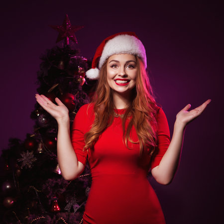 Cute smiling woman in Christmas hat and evening sexy dress. New Year tree with red light on a backgroundの写真素材