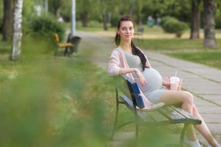 Cute young brunette sexy pregnant woman with long hair in ponytail and in casual dress, walking in european city park. Sunny warm day, outdoor. Copy spaceの写真素材