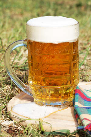glass mug with beer and foam on a wooden tray on the grass on a sunny dayの写真素材