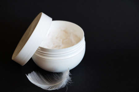 open white jar of cream and white feather on black background, copy space