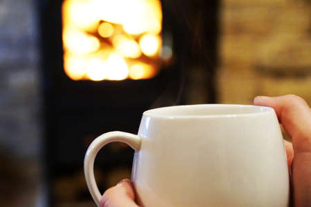 female hands hold a cup of hot tea on the background of a burning fireplace.の写真素材