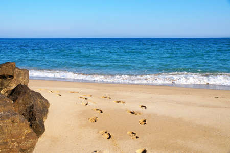 footprints on an empty sandy beach, large stones on the shore, sea horizon and clear sky.の写真素材