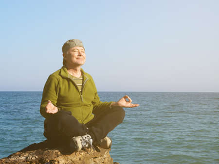 middle aged man sits in lotus position on a large rock by the sea and meditates in sunlight, toned photoの写真素材