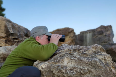 a man leaning on a large stone looks through binoculars.