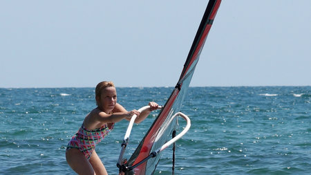 A teenage girl standing on a windsurfing board in the sea tries to hold on to a falling sail with both hands, close-upの写真素材