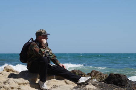A man with a backpack, resting his chin on his hand, looks at the sea horizon while sitting on a large stone on a wild seashore.の写真素材
