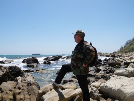 A man with a backpack looks at the sea horizon while standing on a stone on a wild rocky seashore.の写真素材