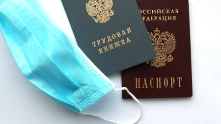 documents with the inscription on the cover "Employment record" and "Russian Federation Passport" and a blue protective mask lying next to it, a symbol of labor relations in a pandemicの写真素材