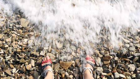 legs of a person in wicker sandals in American flag colors standing on a rocky beach in a spray of...