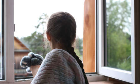 a girl with a pigtail from the back looks out the open window, wrapped in a soft warm knitted blanket and holding a gray plush hare in her handの写真素材