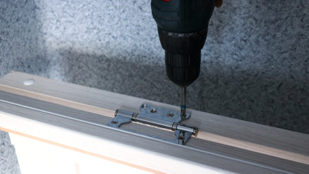 close-up of a drill screwing screws into metal fasteners on a light wooden door frame against a...