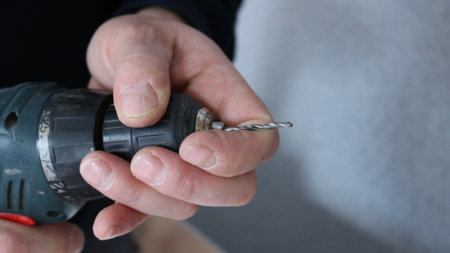 close-up carpenter hands holding green drill body with red button, thin drill setting by male working hands on gray blank wall background with place for textの写真素材
