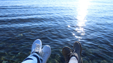 two pairs of legs of people having a rest on the rocky seashore, relaxation of two friends on the sunny seaside, legs in jeans and white sneakers and legs in sweatpants and light boots by the waterの写真素材