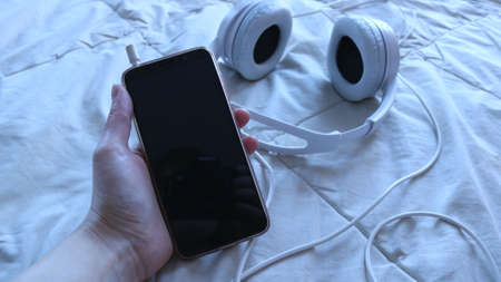 a cell phone with a dark blank screen in a female hand against the background of a soft light blanket on a comfortable bed and white headphones connected to a gadget, copy space on the phone screenの写真素材