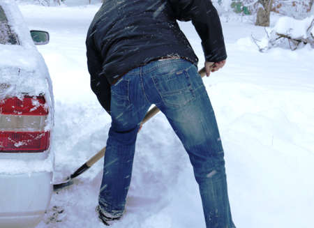a man in jeans and a black jacket clears a place next to a car enter a snowdrift, abnormal snowfalls and their extreme consequences on the roads, shoveling snow and clearing the drivewayの写真素材