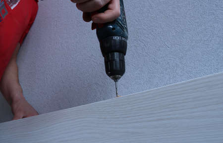 fixing the fasteners on the door leaf of a light wood with a standing end using an electric drill in men's hands, the carpenter drills holes for the fasteners on the door view from the lower angleの写真素材