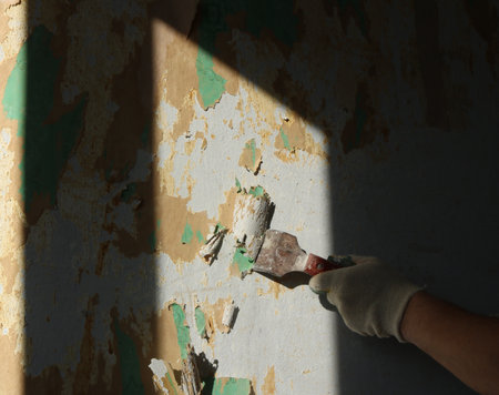 the hand of a worker in a cotton glove with a sharp spatula cuts off scraps of old wallpaper on a sunlit wall in a room, a hand of a builder with a hand tool scrapes off a layer of wall coveringの写真素材