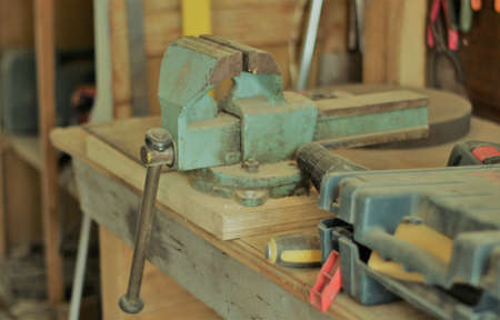 old green iron vise on the workbench of a workshop in a blurry environment of other equipment and tools, a special clamp for parts or products in the workplace of a locksmith or carpenterの写真素材