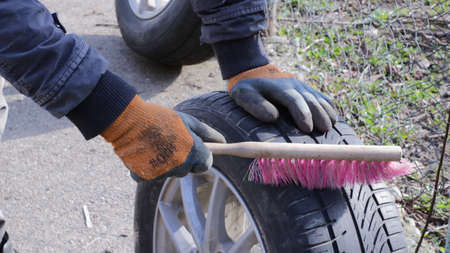 male hands in orange gloves brush dirt from the tire treads on a car wheel, preparing it for installation, home cleaning and seasonal wheel changes outdoorsの写真素材