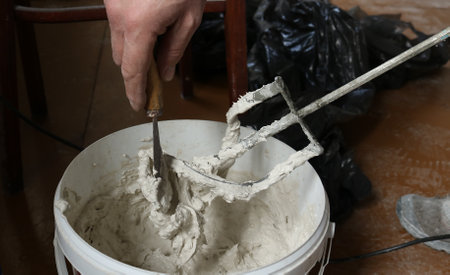 scraping with a small spatula the mortar from a construction mixer over a container, the hands of a plasterer working with plastic mastic in a white bucket, mixing the putty with a large whiskの写真素材