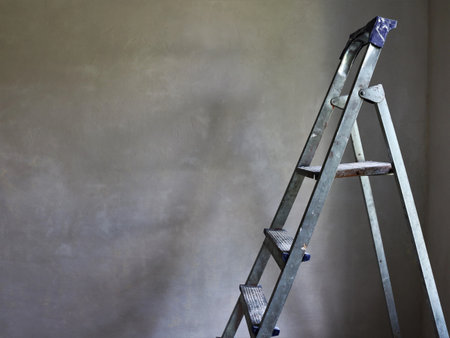 the top of a folding aluminum ladder visible against the background of an empty gray freshly plastered wall as a vintage background on the theme of renovation with space for textの写真素材