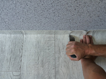 trimming linoleum with a light wood texture along the junction line with a wall with gray abstract wallpaper with a sharp short knife in male hands, fitting the floor coveringの写真素材