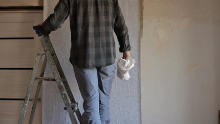 a man in gray clothes stands on an aluminum stepladder with a white rag in his hand and sticks plain wallpaper on a prepared wall, do-it-yourself wallpapering in a houseの写真素材