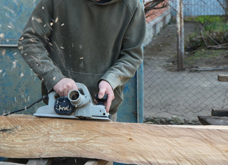 carpenter with electric plane sanding surface of wooden material in outdoor workshop with flying...