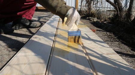 An employee wearing work clothes and gloves uses a brush to apply yellow paint or varnish to light-colored wooden boards that lie on the pavement.の写真素材