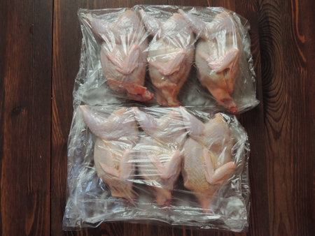 skinned domestic quail carcasses wrapped in three plastic bags placed on a textured dark stained wood countertop, farm-raised featherless poultry in clear packaging materialの写真素材