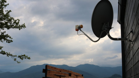 silhouette of satellite dish hanging on wooden wall of house or cottage against beautiful cloudy sky and mountain ranges, communication and connectivity in remote places for travelの写真素材