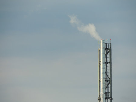 high metal chimney of autonomous boiler station with smoke clouds and built-in ladder against light...