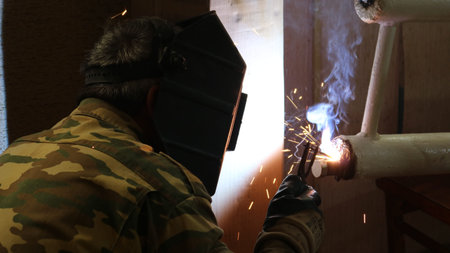A welder, seen from behind, in uniform and a protective helmet, welds a section of a home heating...