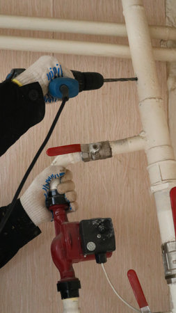 A repairman's gloved hands hold a drill and drill a hole in a white iron pipe in the structure of a...