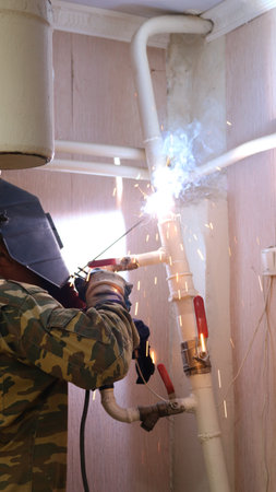A welder, seen from behind, wearing a uniform and hard hat, welds a section of a house's heating...