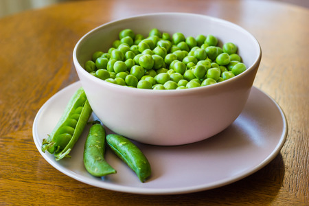 pink bowl with green peas and three pea pods lie near stands on a wooden table close-up shallow depth of fieldの写真素材