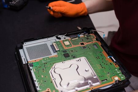 SAINT-PETERSBURG, RUSSIA, OCTOBER 27 2019: Man in orange gloves parses the PlayStation 4 Pro console with a screwdriver. Close-up, shallow depth of fieldのeditorial素材