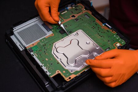SAINT-PETERSBURG, RUSSIA, OCTOBER 27 2019: Man in orange gloves parses the PlayStation 4 Pro console with tweezers. Close-up, shallow depth of fieldのeditorial素材