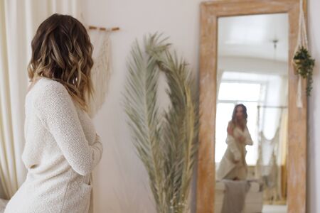 Blonde European female wearing tender fashionable cardigan sitting in bright modern bedroom looking at big mirror. Picture with copy space, selective focus. quarantine, stay homeの写真素材