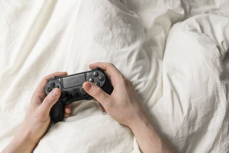 Gamer man sits in rumpled bed with wireless gamepad in his hands. Stay home due quarantine. Leisure...