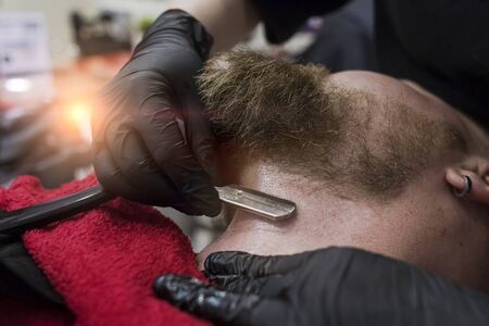 Shaving a beard and neck with a straight razor. Haircut in fashionable barbershop. Hands of barber in black gloves shave a client. Close-up shallow depth of field.の写真素材